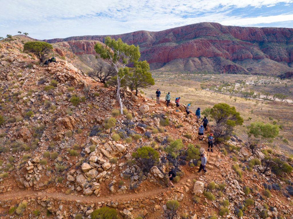 Group of hikers trekking on rocky trail through Australian outback with red mountain ranges in background