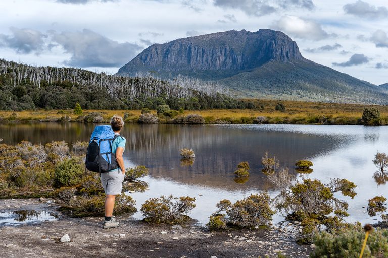 Hiker with blue backpack viewing Mount Oakleigh across alpine lake in Tasmania's wilderness