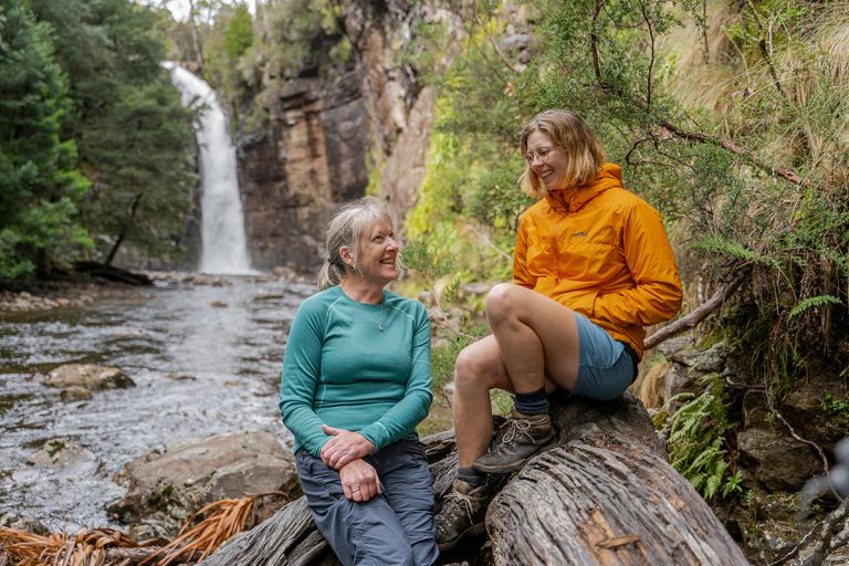Two hikers resting on a log near a waterfall in a lush forest setting wearing outdoor gear