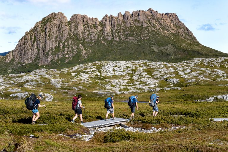 Hikers with backpacks crossing wooden boardwalk on moorland with dramatic rocky mountain peak in background